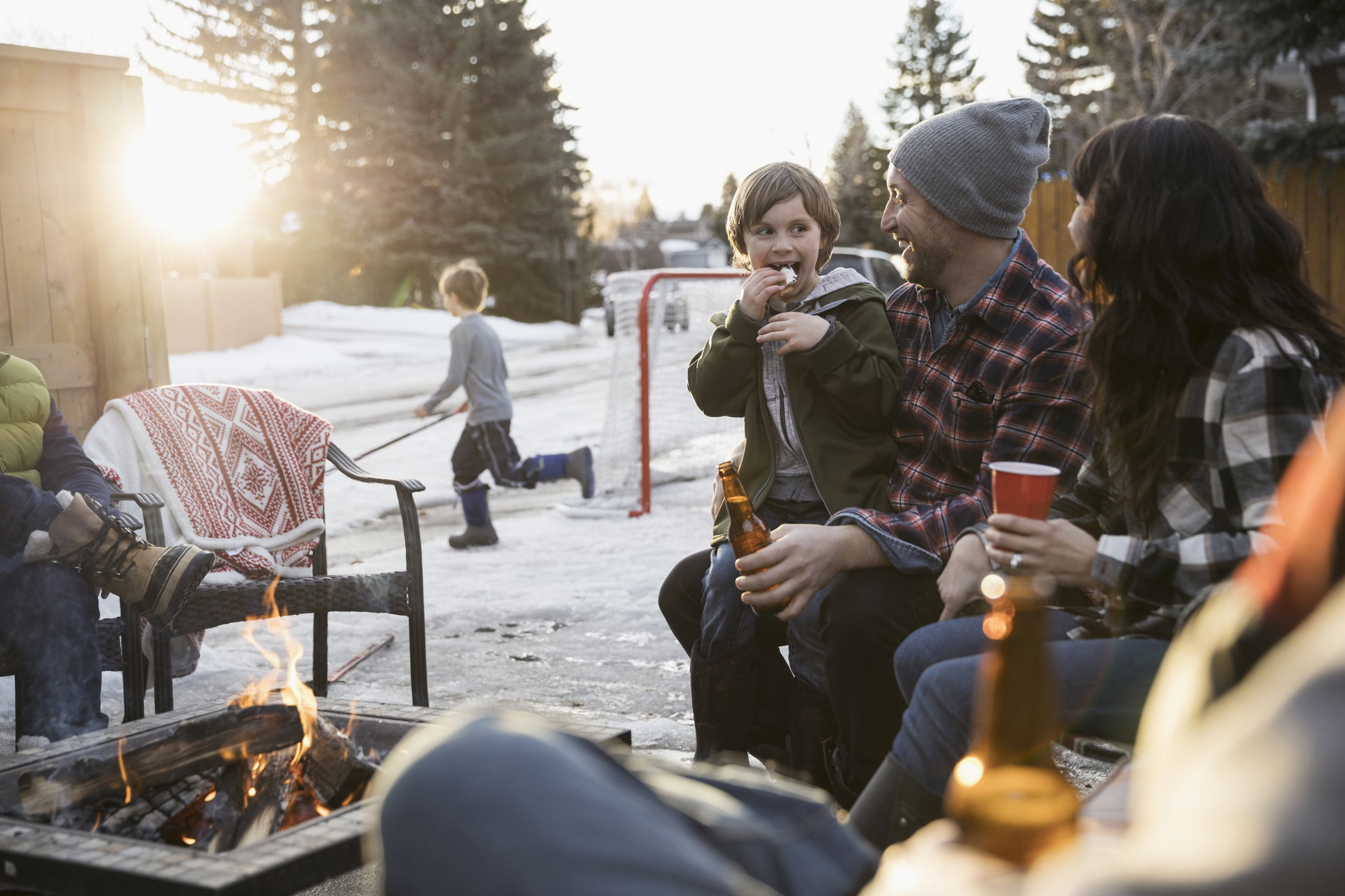 Family roasting marshmallows and drinking beer at fire pit in snowy driveway