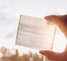 Person holding natural mineral stone Selenite plate against sun and blue sky, stone has healing and cleansing properties. Also used for recharging other mineral crystal stones.