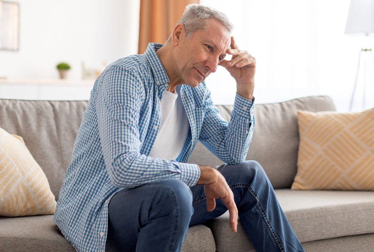 mężczyzna w wieku 60 plus w jasnej koszuli w kratkę siedzi na kanapie zmartwiony i rozmyśla