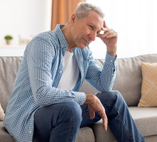 mężczyzna w wieku 60 plus w jasnej koszuli w kratkę siedzi na kanapie zmartwiony i rozmyśla