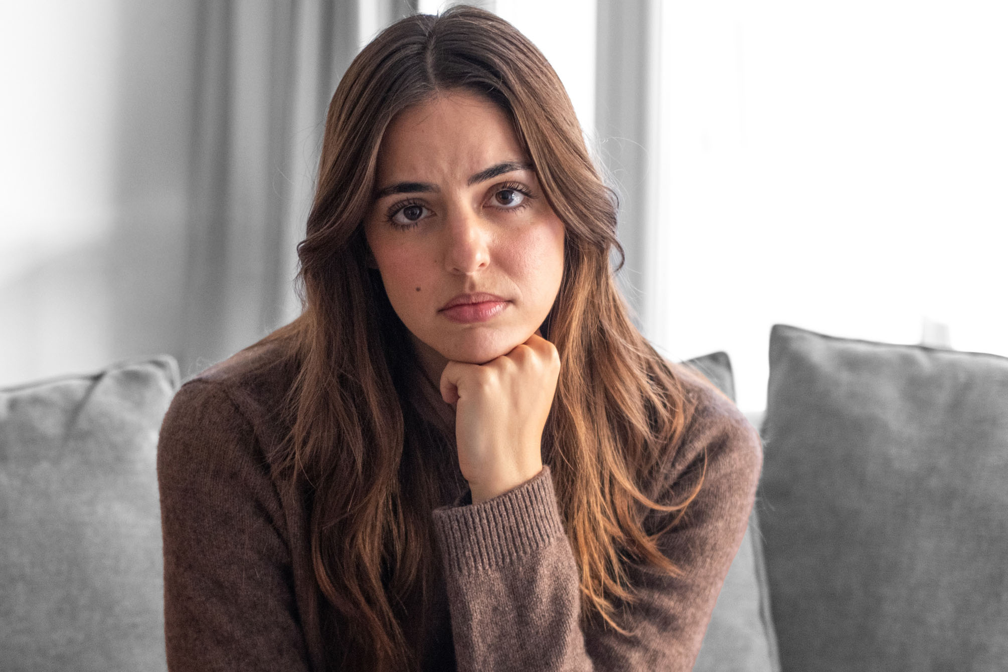Portrait of serious and pensive young woman with one hand under the chin sitting on couch at home looking at camera.