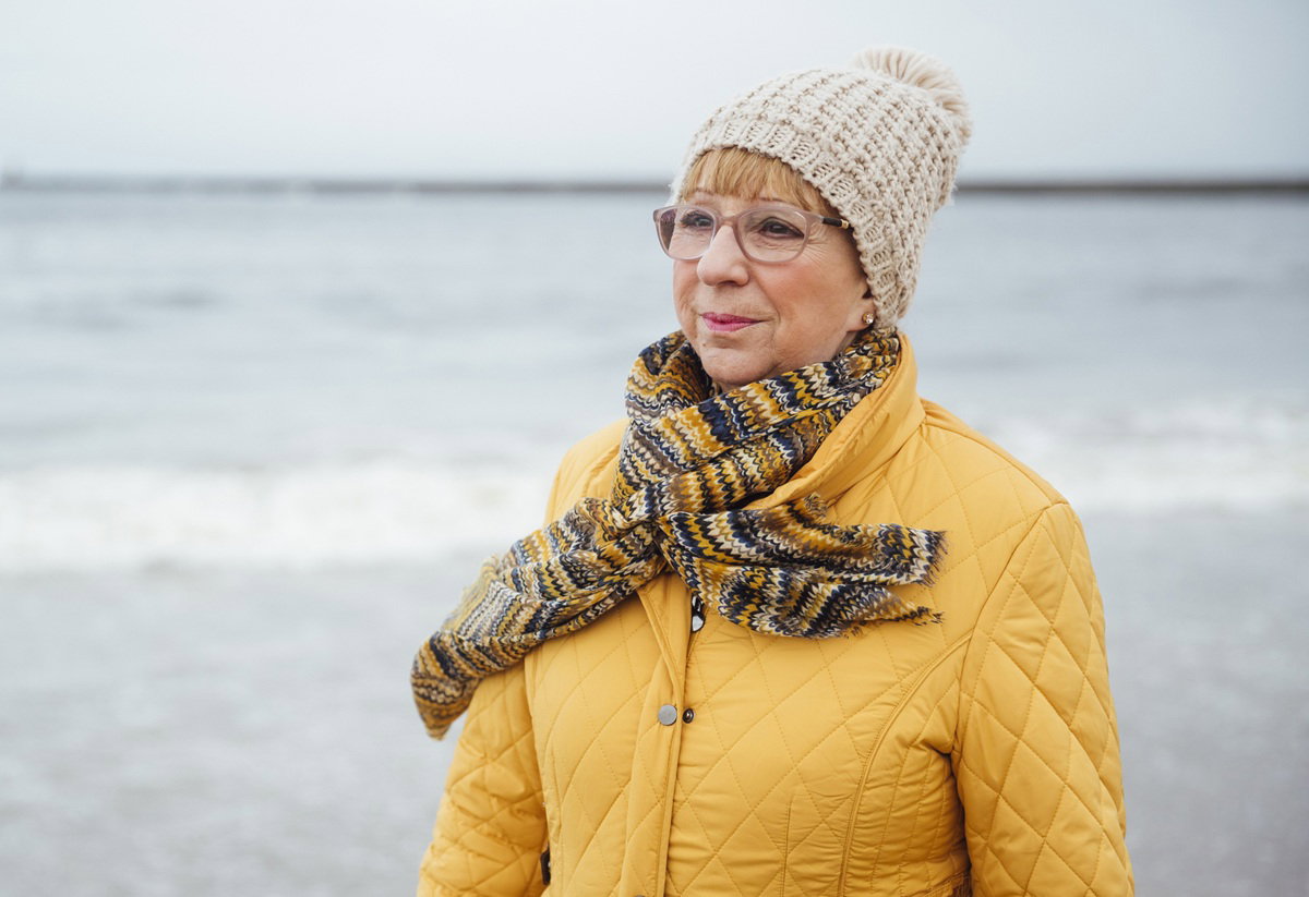 kobieta w wieku 65 plus w kurtce i w okularach stoi na tle morza zamyślona