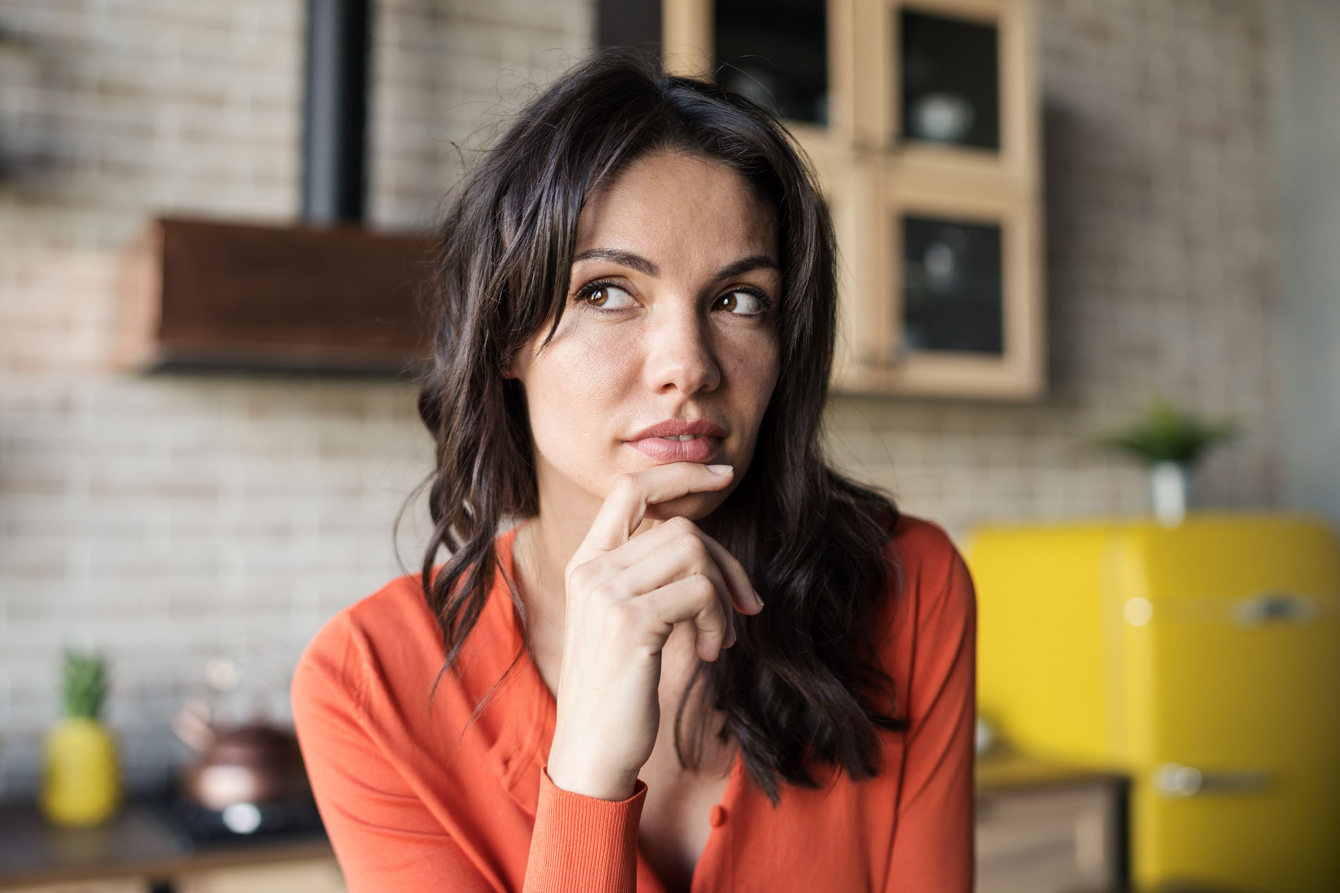 Portrait of beautiful brunette woman thinking or creating idea with her hand on chin in kitchen at home