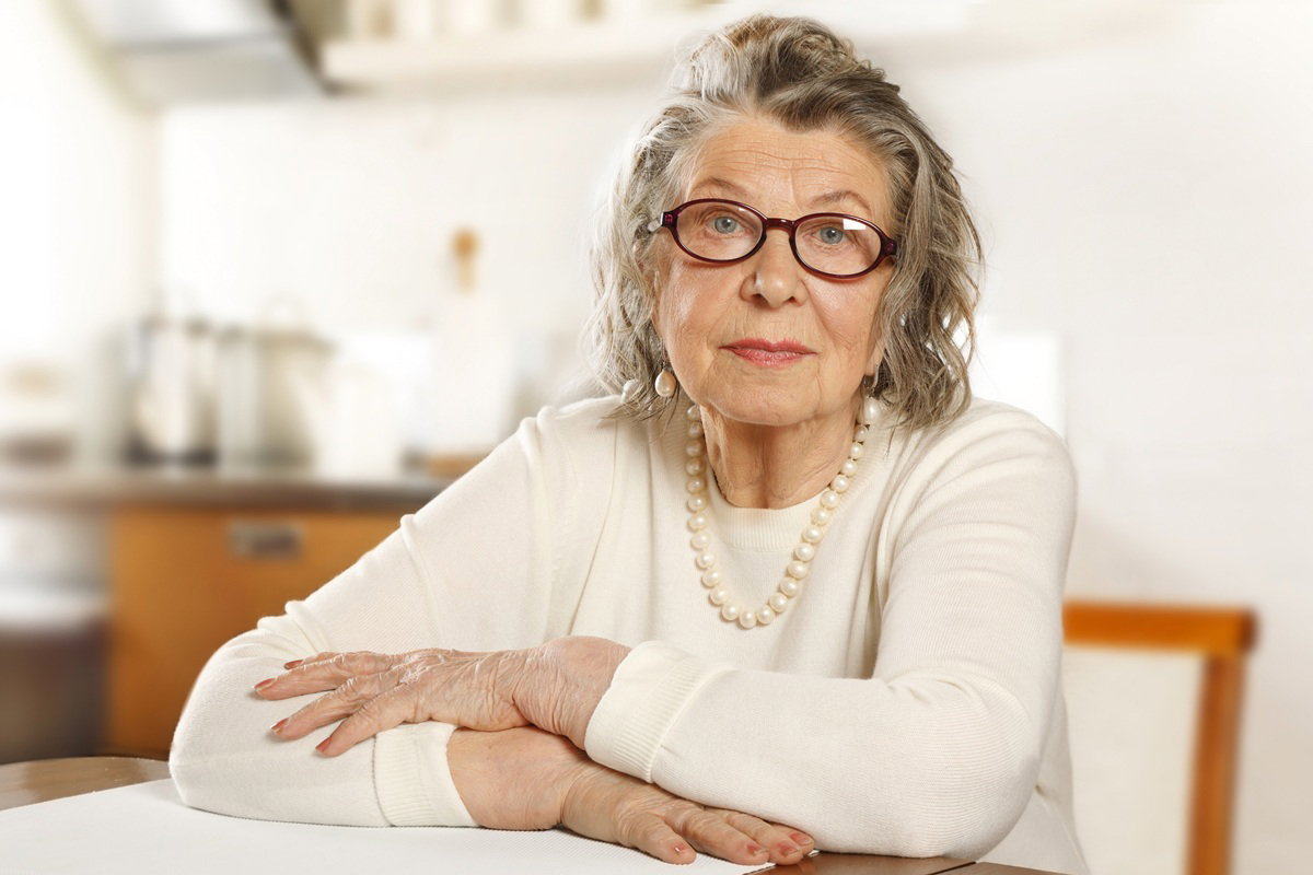 kobieta w wieku 70 plus w okularach z perłami na szyi siedzi smutna przy stole