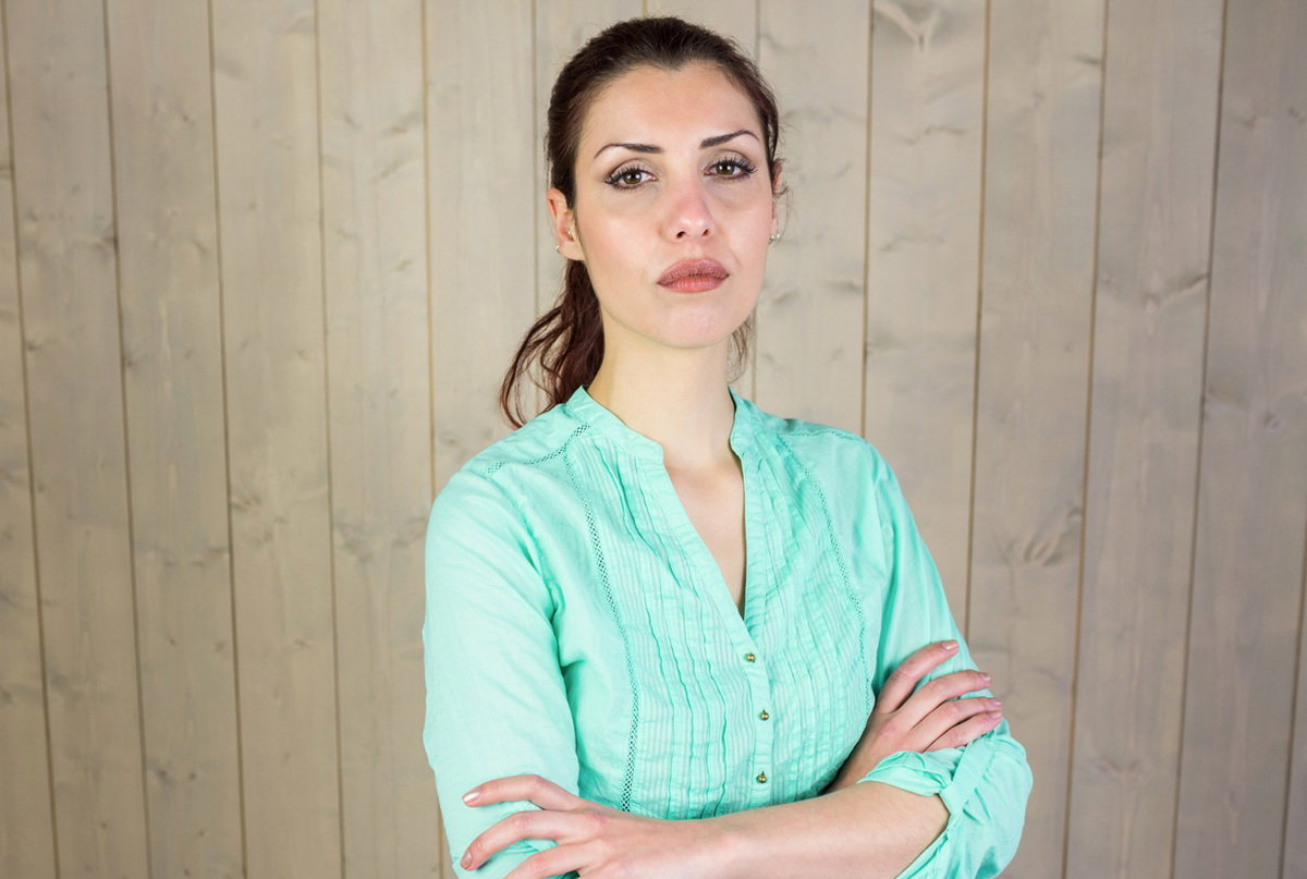 kobieta w wieku 25 plus w zielonej bluzce na tle drewnianej ściany z zaciętą miną