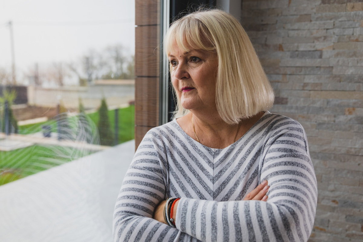 kobieta w wieku 60 plus blondynka w bluzce w paski stoi przy oknie zamyślona i smutna