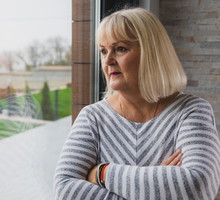 kobieta w wieku 60 plus blondynka w bluzce w paski stoi przy oknie zamyślona i smutna