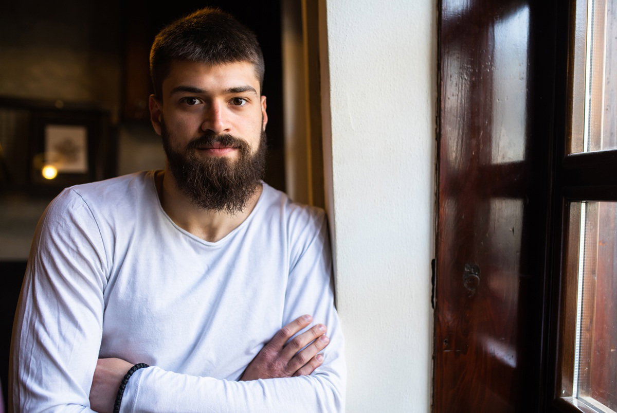 mężczyzna w wieku 30 plus z brodą i w białym longsleevie stoi zamyślony