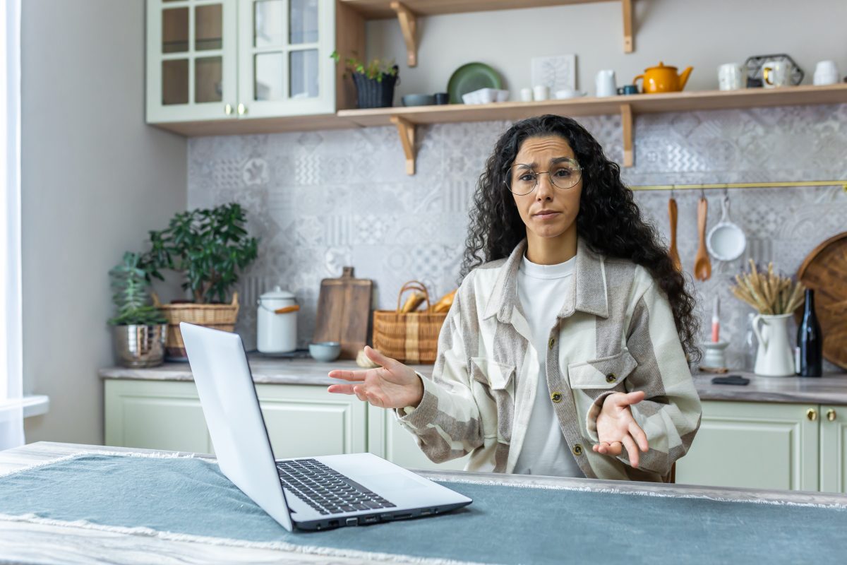 Portrait of unhappy woman with laptop at home, hispanic woman looking at camera disappointed in kitchen with curly hair and glasses