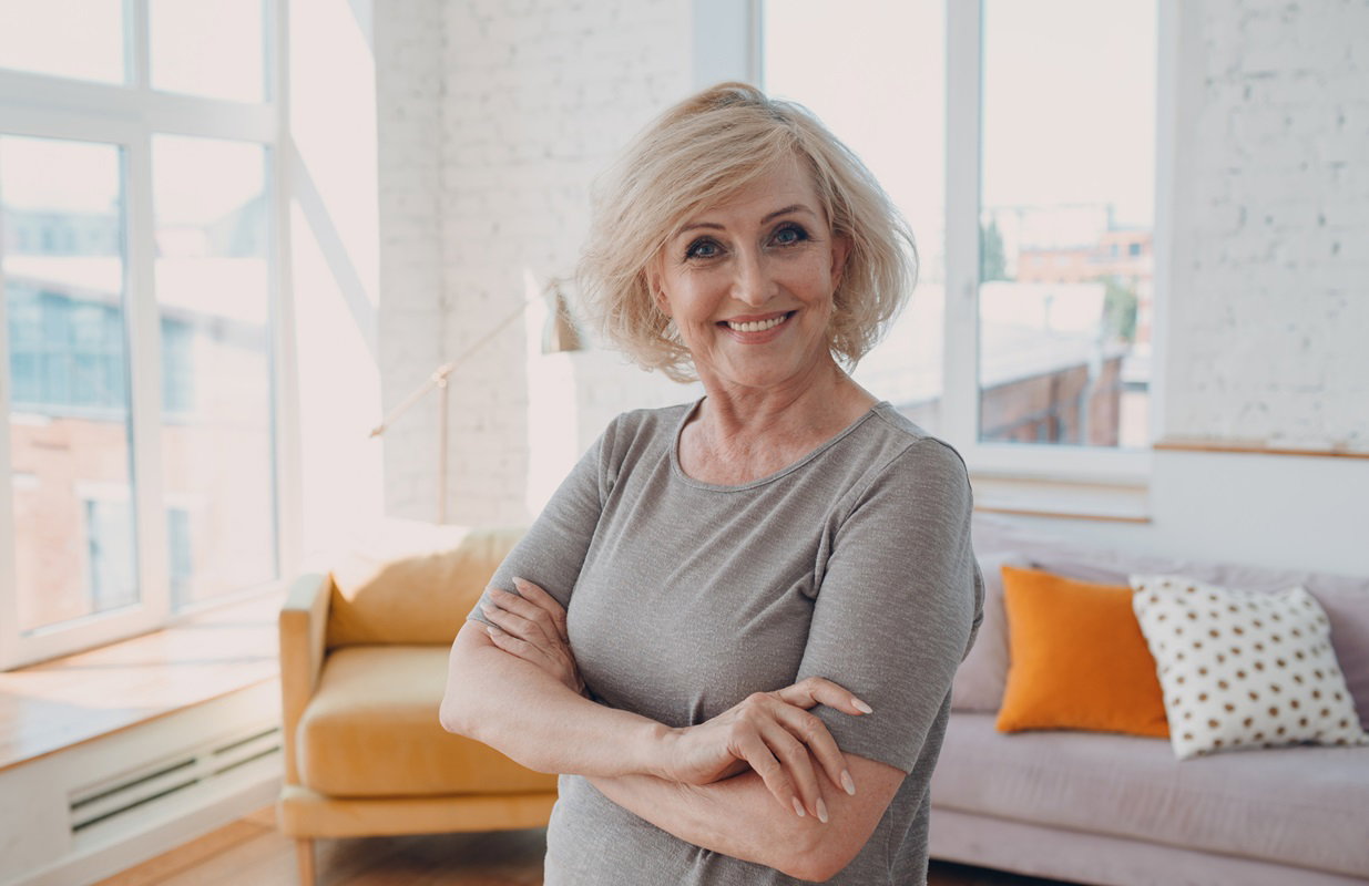 seniorka blondynka z szerokim uśmiechem stoi z założonymi rękami w salonie