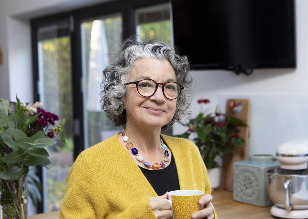 kobieta w żółtym swetrze w senioralnym wieku z kręconymi włosami i w okularach stoi z kubkiem w ręku i się uśmiecha