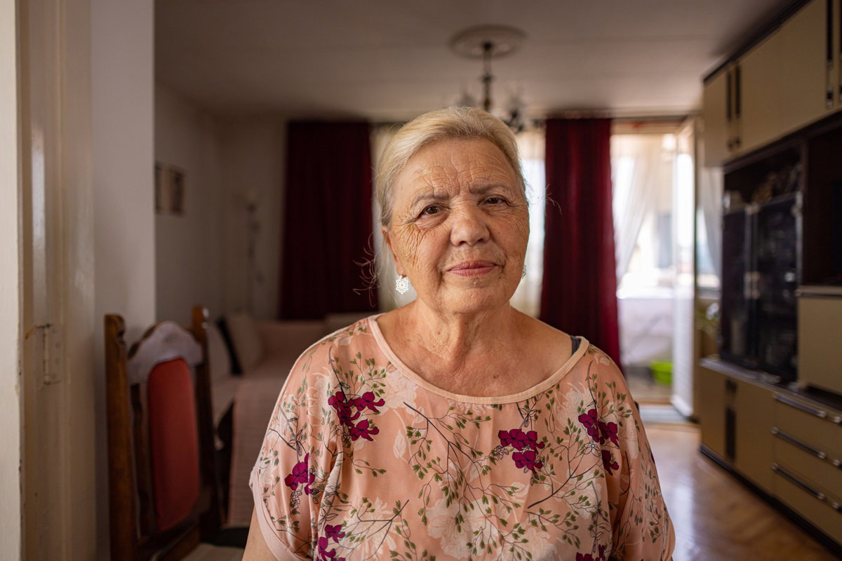 starsza kobieta w kwiecistej bluzce stoi na środku salonu ze zmartwioną miną
