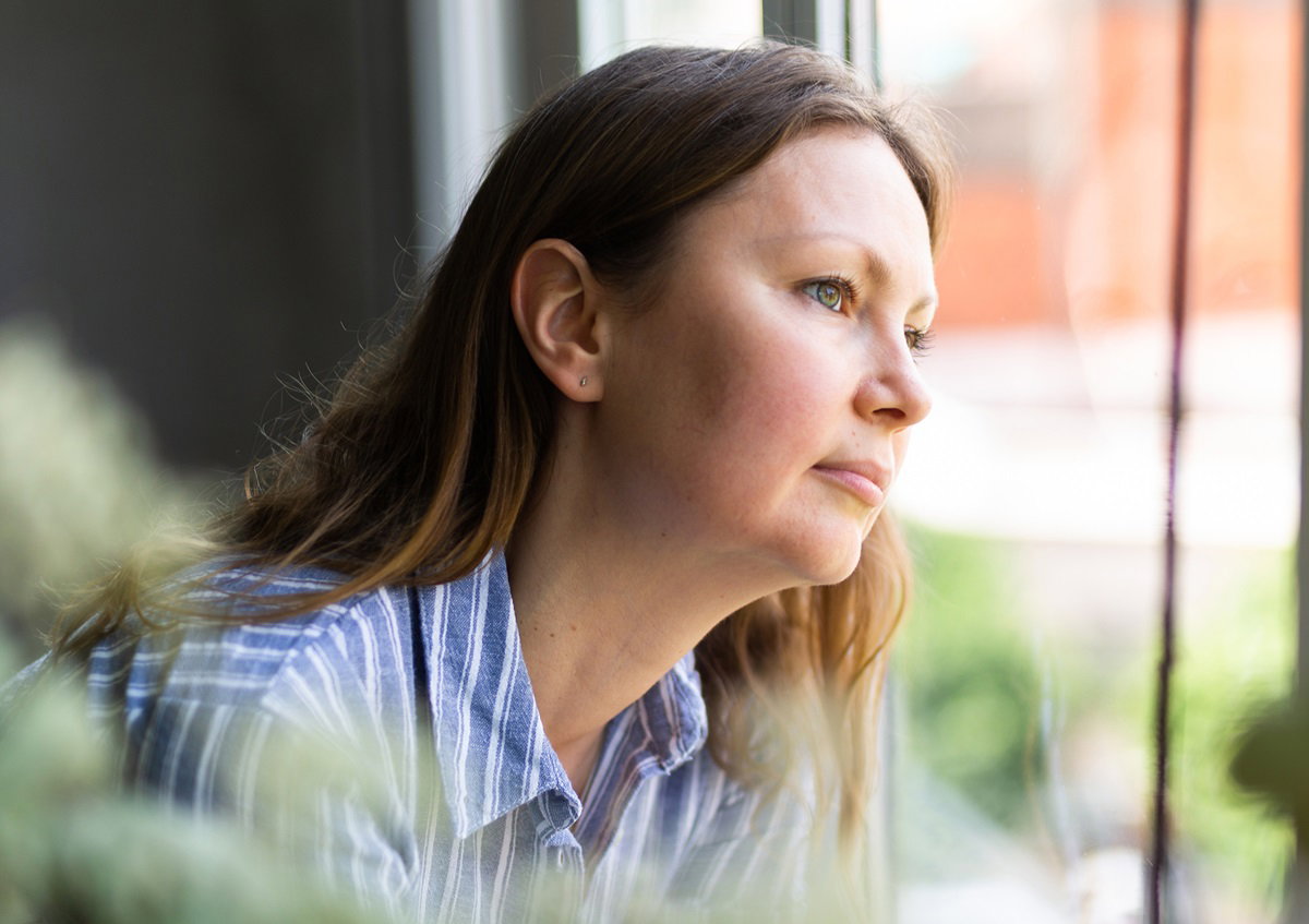 kobieta w średnim wieku z długimi włosami wygląda przez okno i ma smutny wzrok
