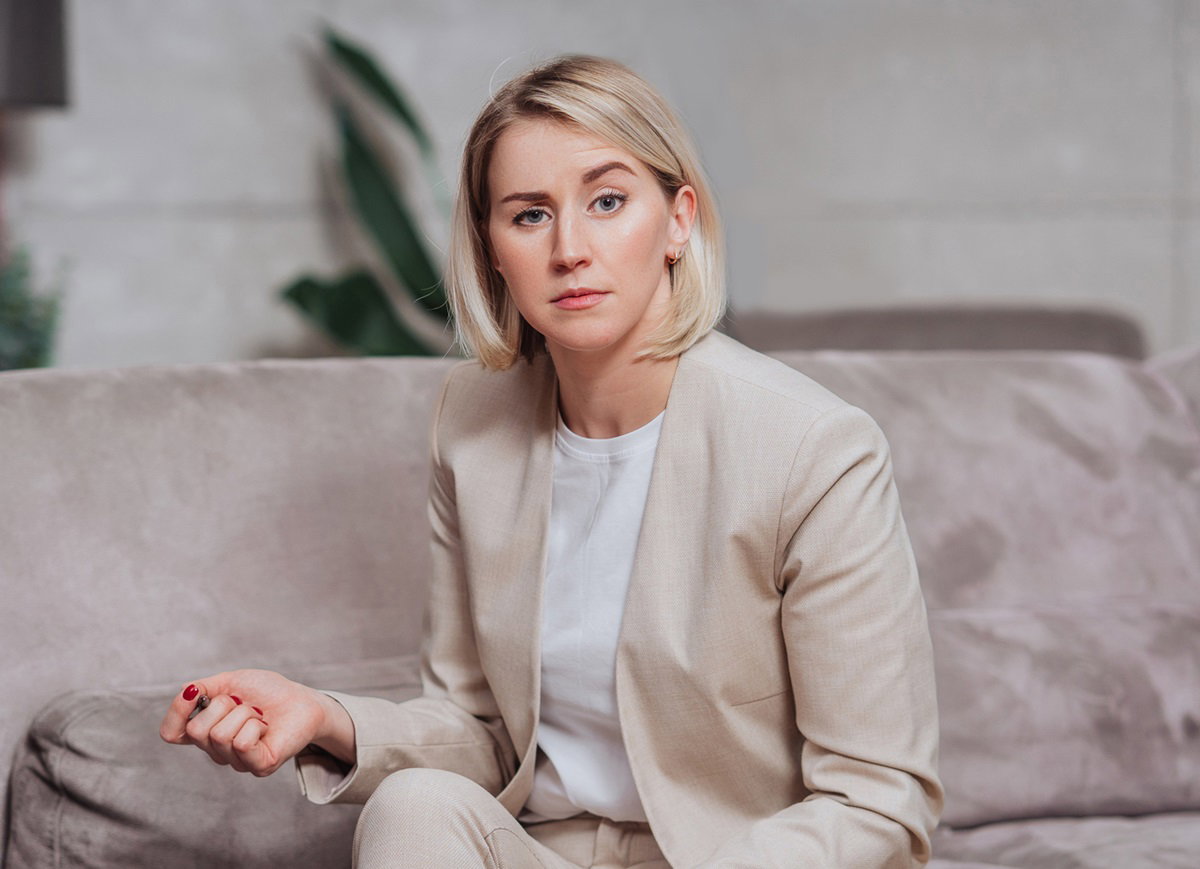 młoda kobieta z blond włosami  w beżowym kostiumie siedzi na kanapie ze zdziwioną miną