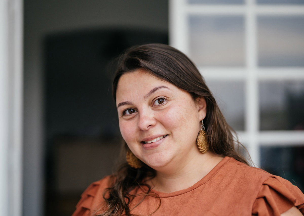 młoda kobieta z ciemnymi włosami i z kolczykami z lekkim uśmiechem na tle domu