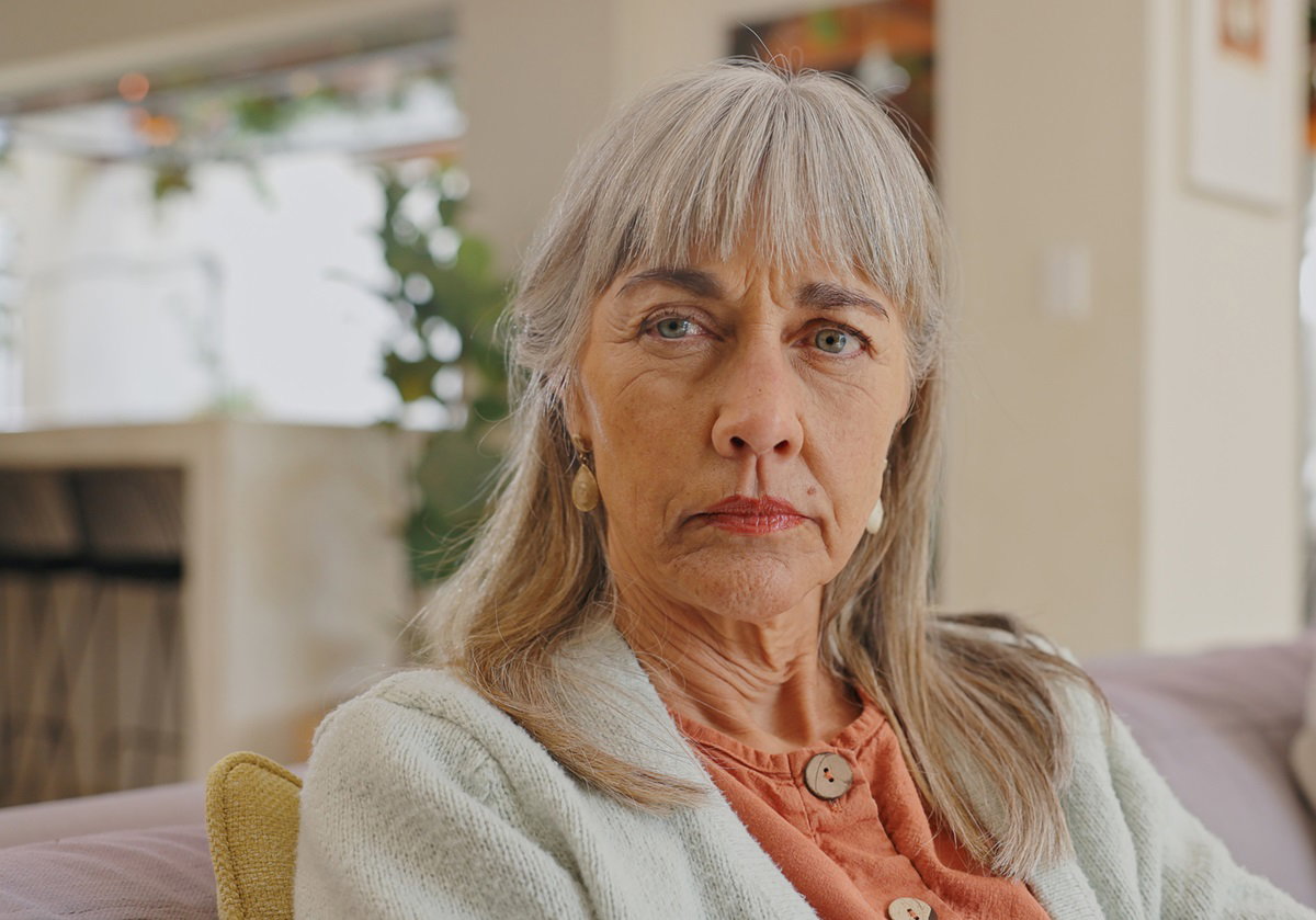 kobieta w starszym wieku z długimi włosami i grzywką siedzi na kanapie z groźną miną