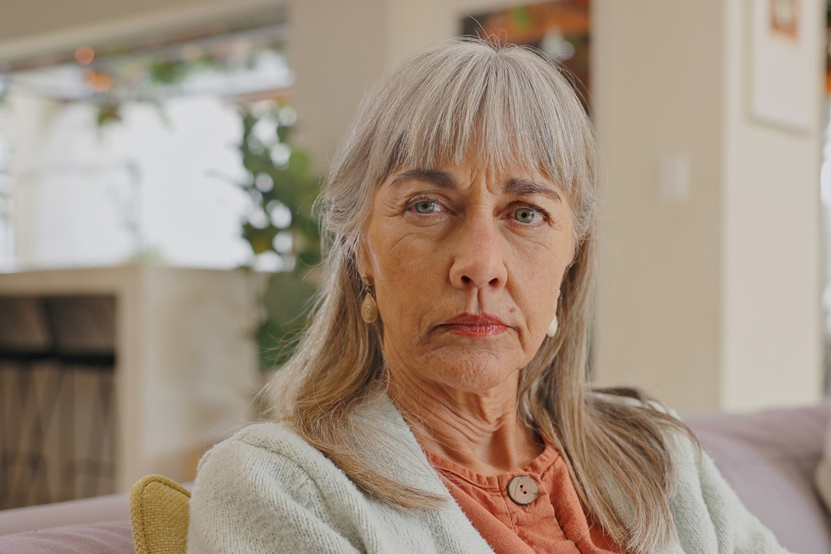 kobieta w starszym wieku z długimi włosami i grzywką siedzi na kanapie z groźną miną