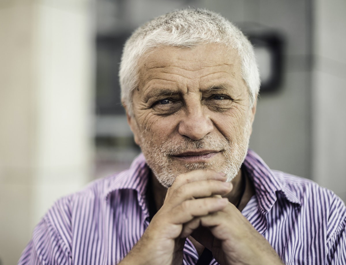 starszy mężczyzna z siwymi włosami patrzy przed siebie i ma splecione ręce jak do modlitwy