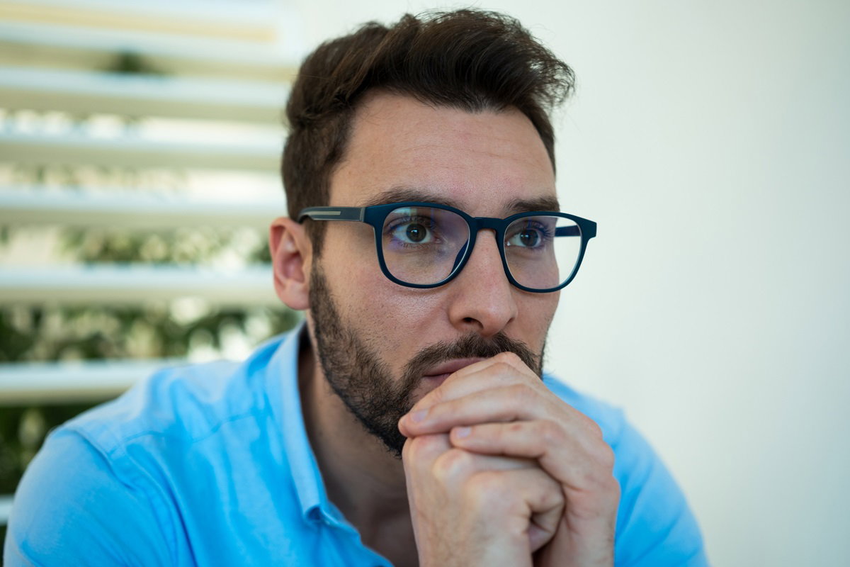 młody mężczyzna w okularach siedzi zmartwiony z rękami przy ustach