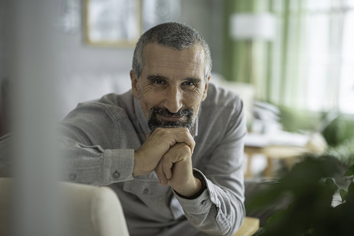 dojrzały mężczyzna w szarej koszuli siedzi na kanapie i patrzy przed siebie z uśmiechem