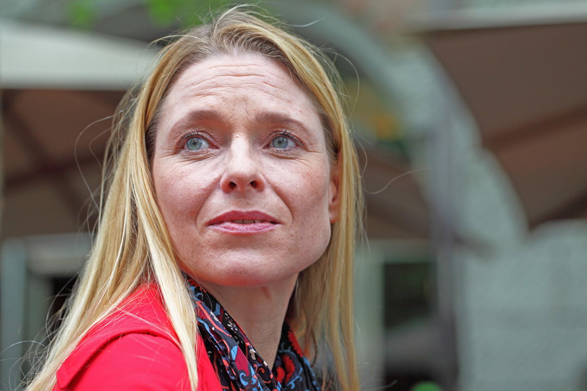 kobieta w średnim wieku z długimi blond włosami i w czerwonej bluzce patrzy w dal zrozpaczona