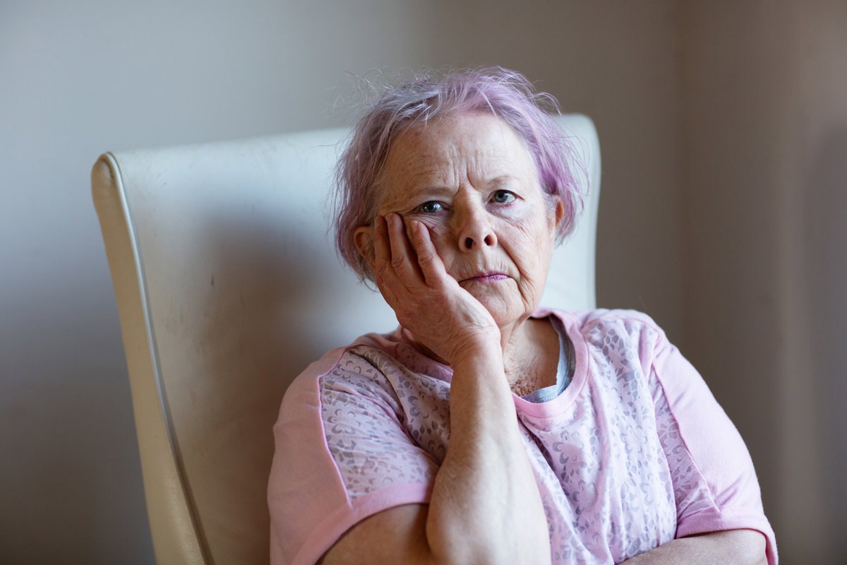 starsza kobieta z włosami pofarbowanymi na fioletowo siedzi w fotelu ze zmartwioną miną