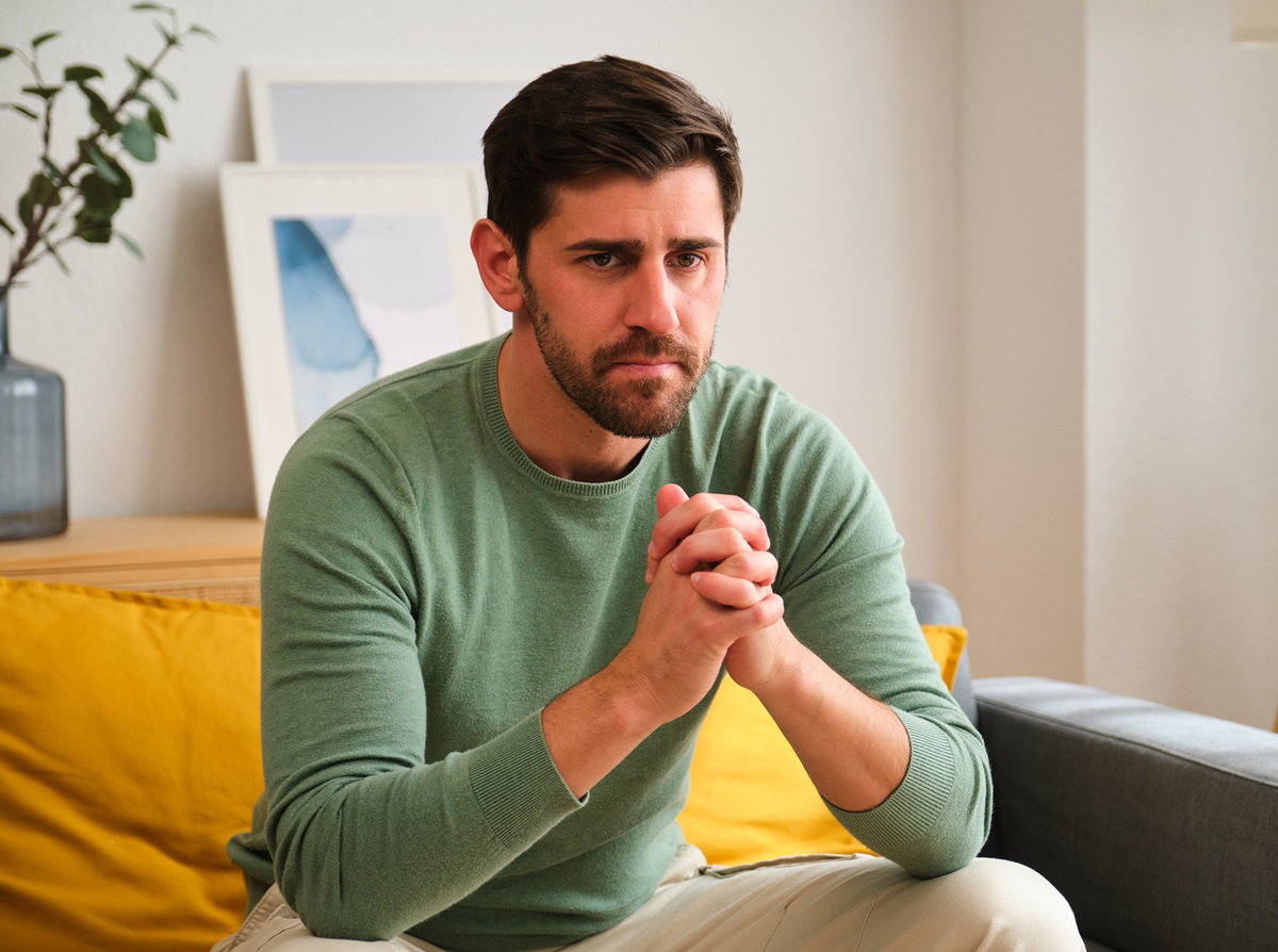 mężczyzna  około czterdziestki w zielonym swetrze siedzi na kanapie w salonie ze zmartwioną miną i złożonymi rękami