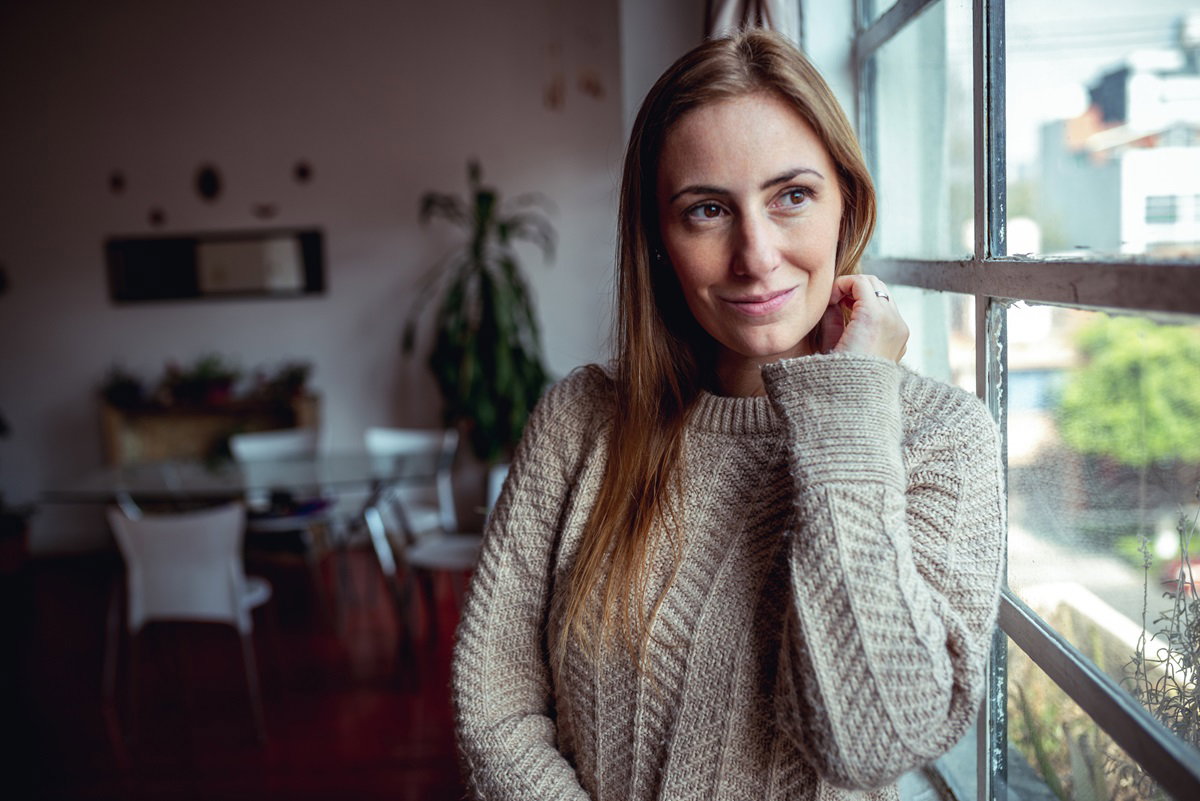 młoda kobieta z długimi włosami i w beżowym swetrze stoi i patrzy w okno rozmarzonym wzrokiem