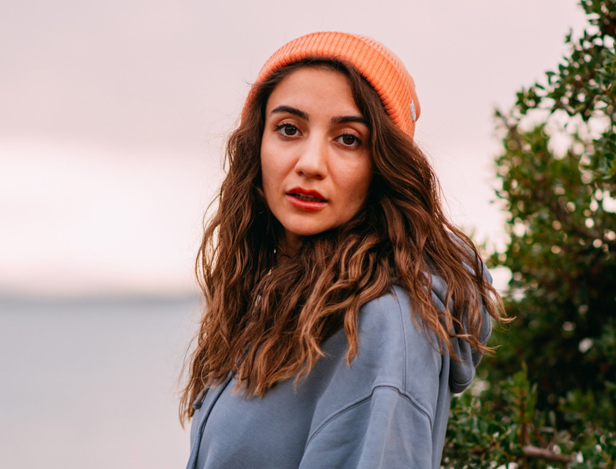 młoda kobieta z długimi pofalowanymi włosami i w pomarańczowej czapce stoi nad brzegiem jeziora ze smutną miną