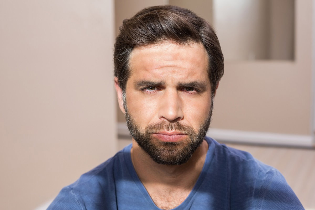 mężczyzna w średnik wieku w niebieskim t-shircie i z brodą siedzi i parzy przed siebie ze zmartwiona miną