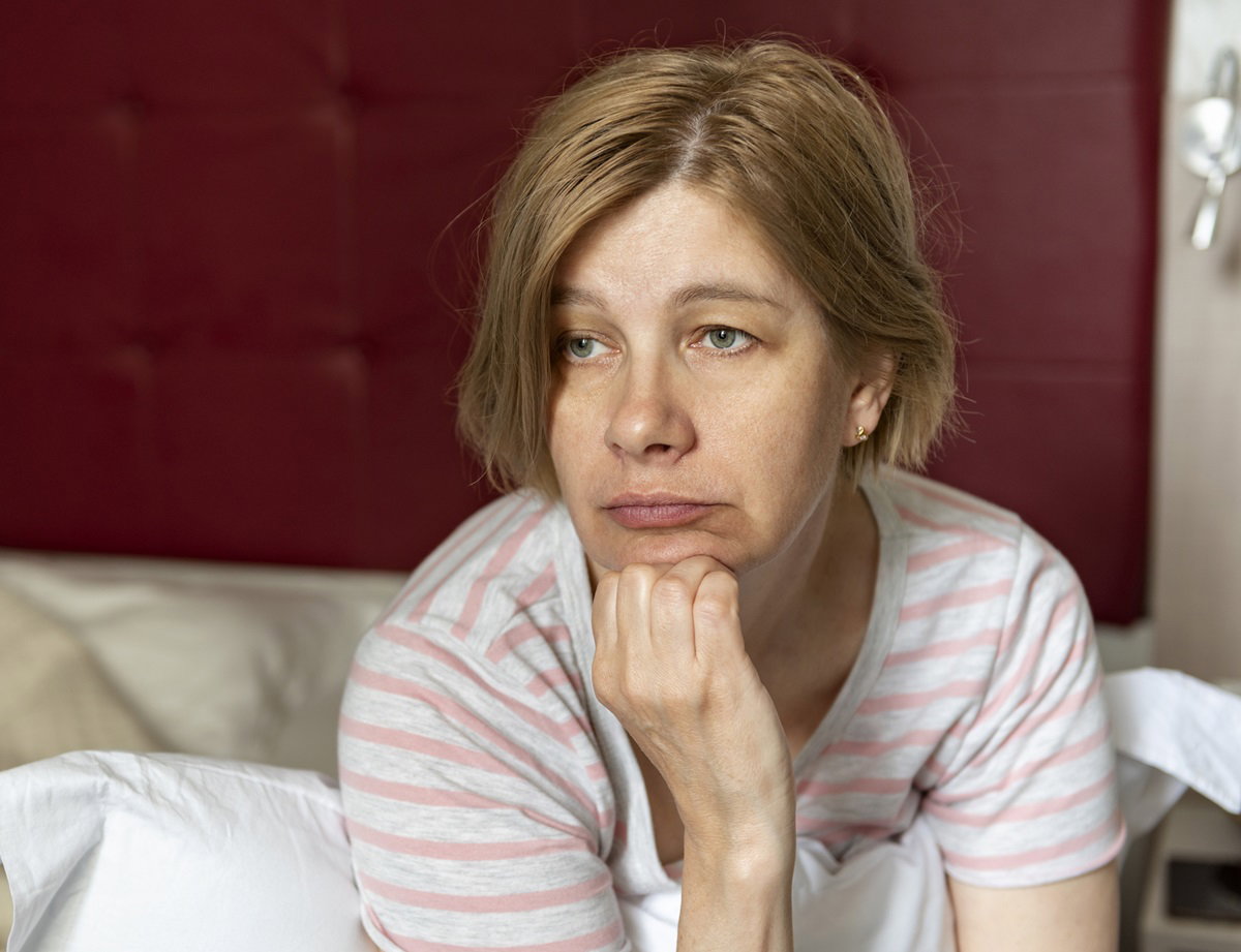 kobieta w średnim wieku siedzi w sypialni na łóżku i patrzy zmartwiony wzrokiem przed siebie