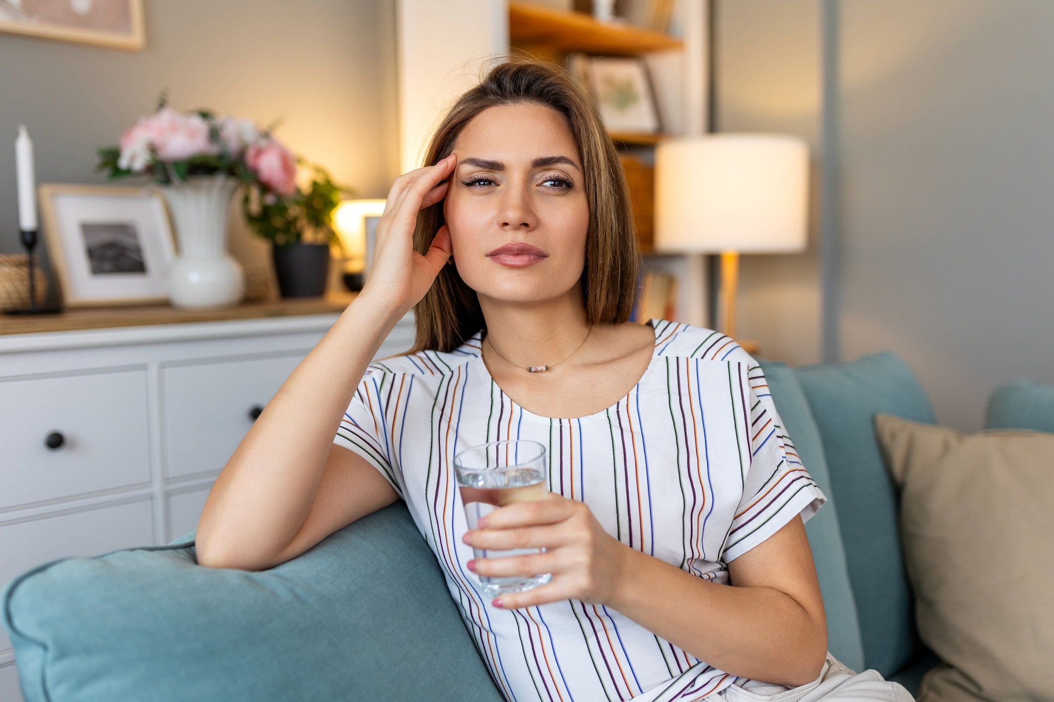 Portrait of an attractive woman sitting on a sofa at home with a headache, feeling pain and with an expression of being unwell.