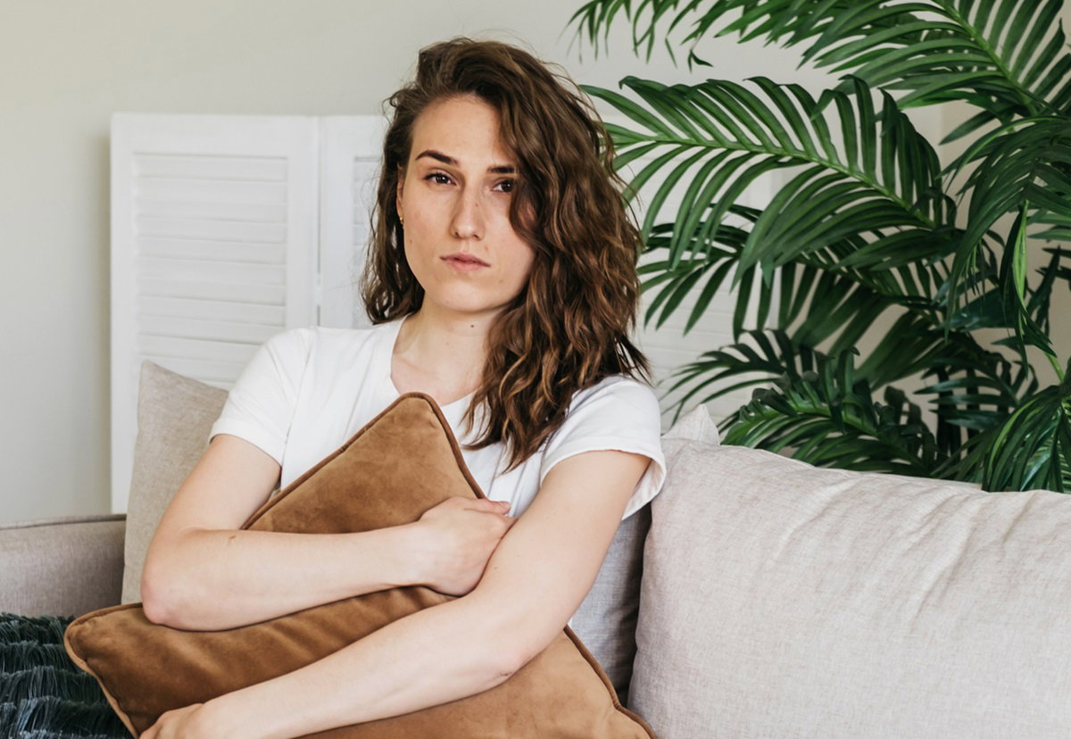 młoda kobieta  z długimi pofalowanymi włosami siedzi na kanapie, ściskając poduszkę i patrzy zranionym spojrzeniem