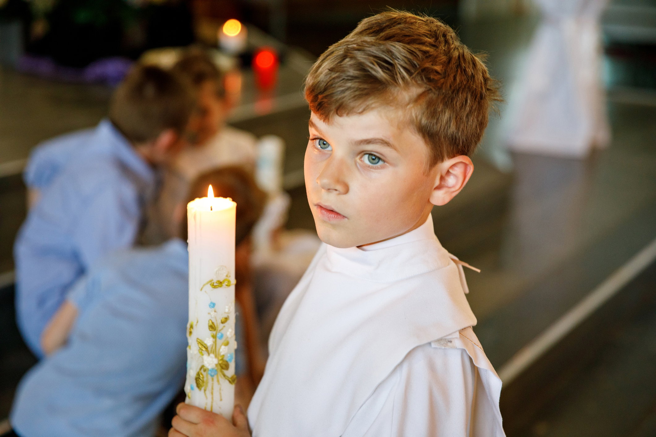 Little kid boy receiving his first holy communion. Happy child holding Christening candle. Tradition in catholic curch. Kid in a white traditional gown in a church near altar.