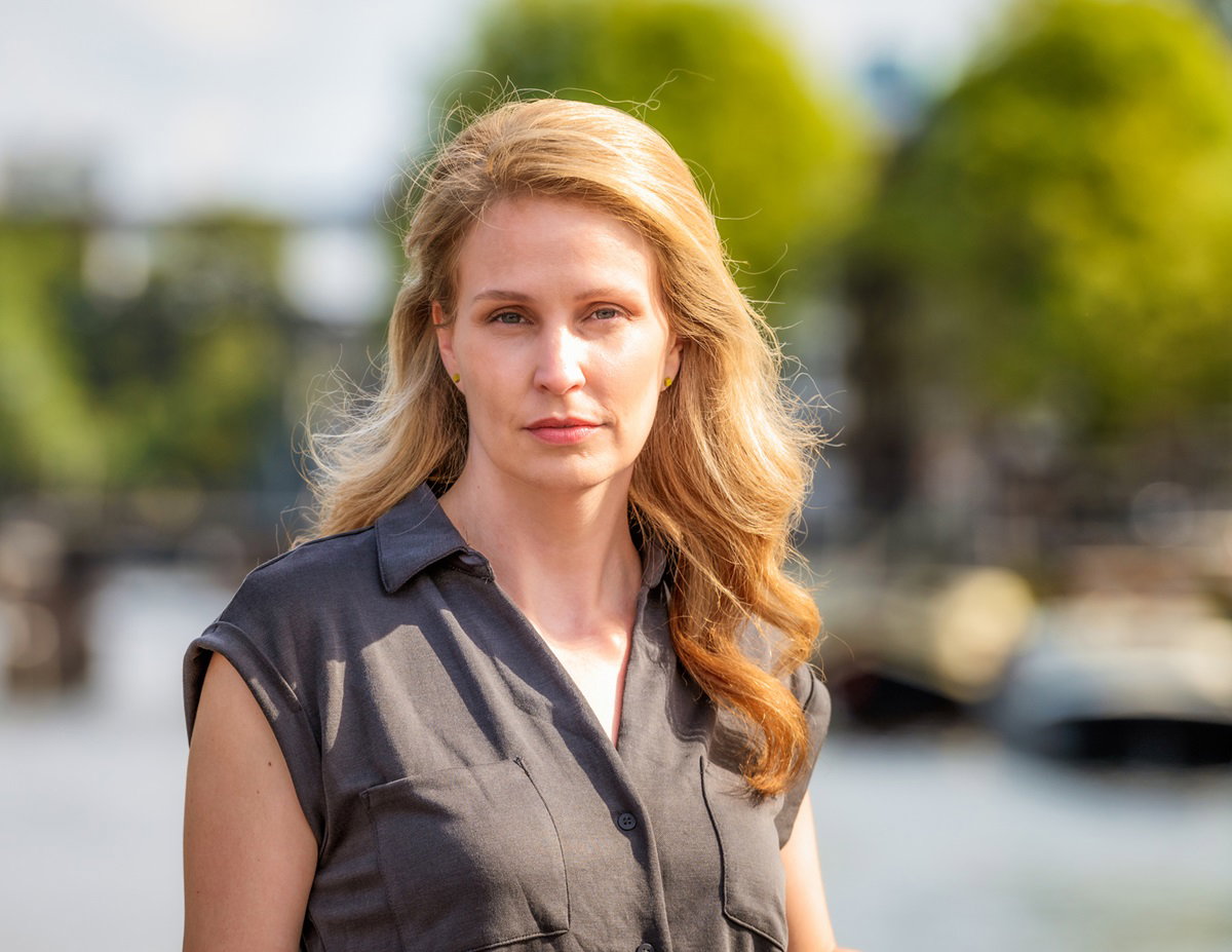 kobieta z długimi blond włosami i brązowej bluzce stoi i patrzy zamyślona