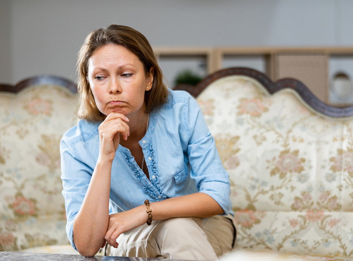 kobieta w średnim wieku w błękitnej bluzce siedzi na sofie i ma zmartwioną minę
