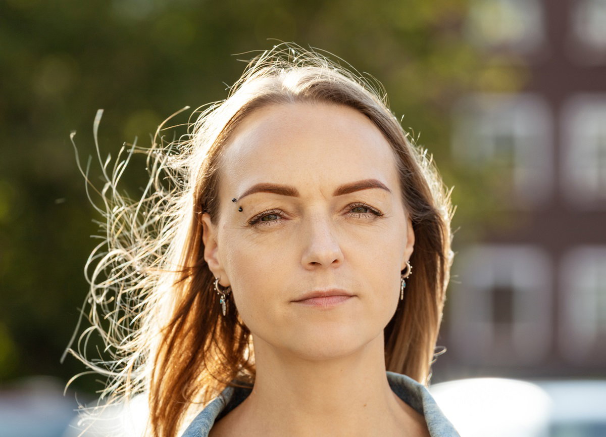 kobieta w średnim wieku z kolczykiem w brwi i jasnymi włosami stoi na ulicy i patrzy poważnym wzrokiem