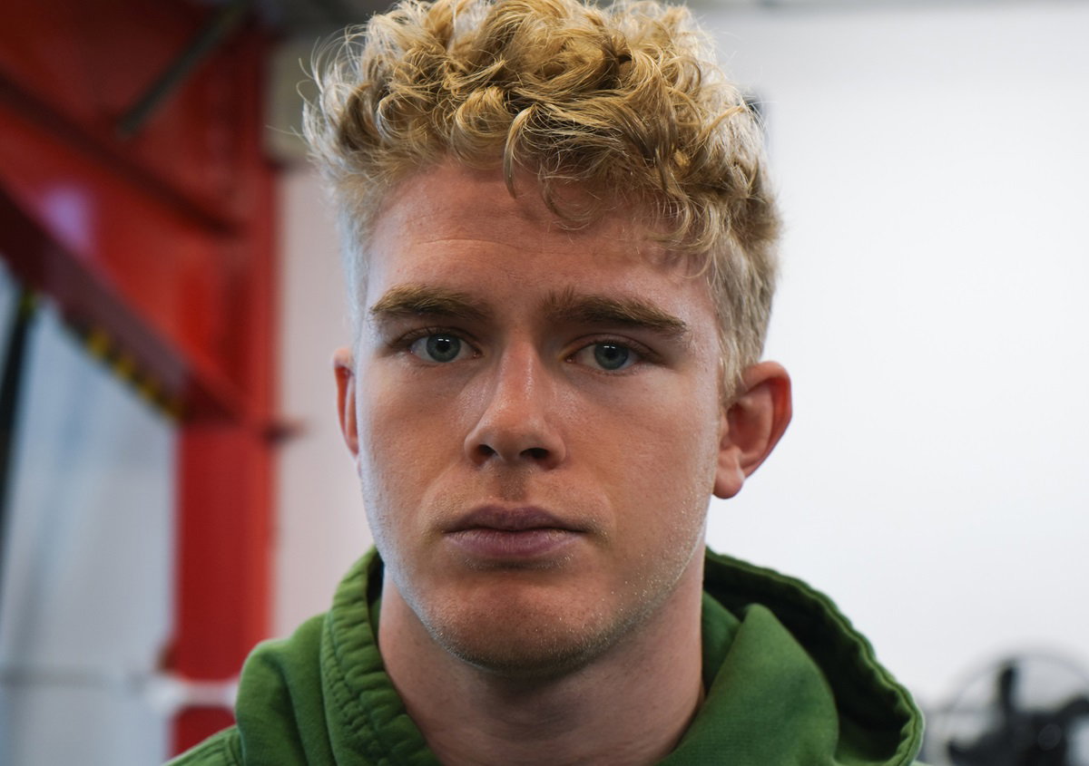 młody chłopak z burzą blond loków w zielonej bluzie patrzy przed siebie smutnym wzrokiem