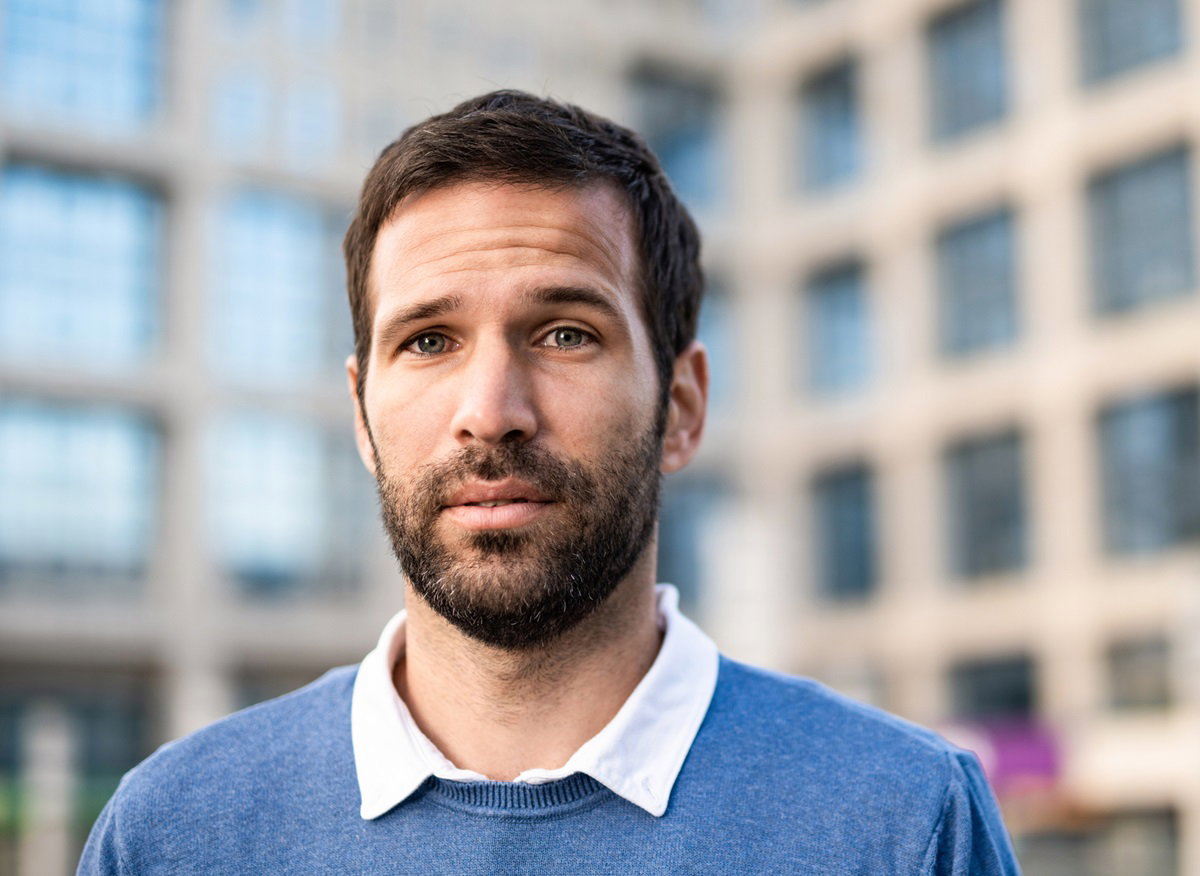 młody mężczyzna w sweterku i z kołnierzykiem stoi na tle biura w wieżowcu i ma zmartwioną minę