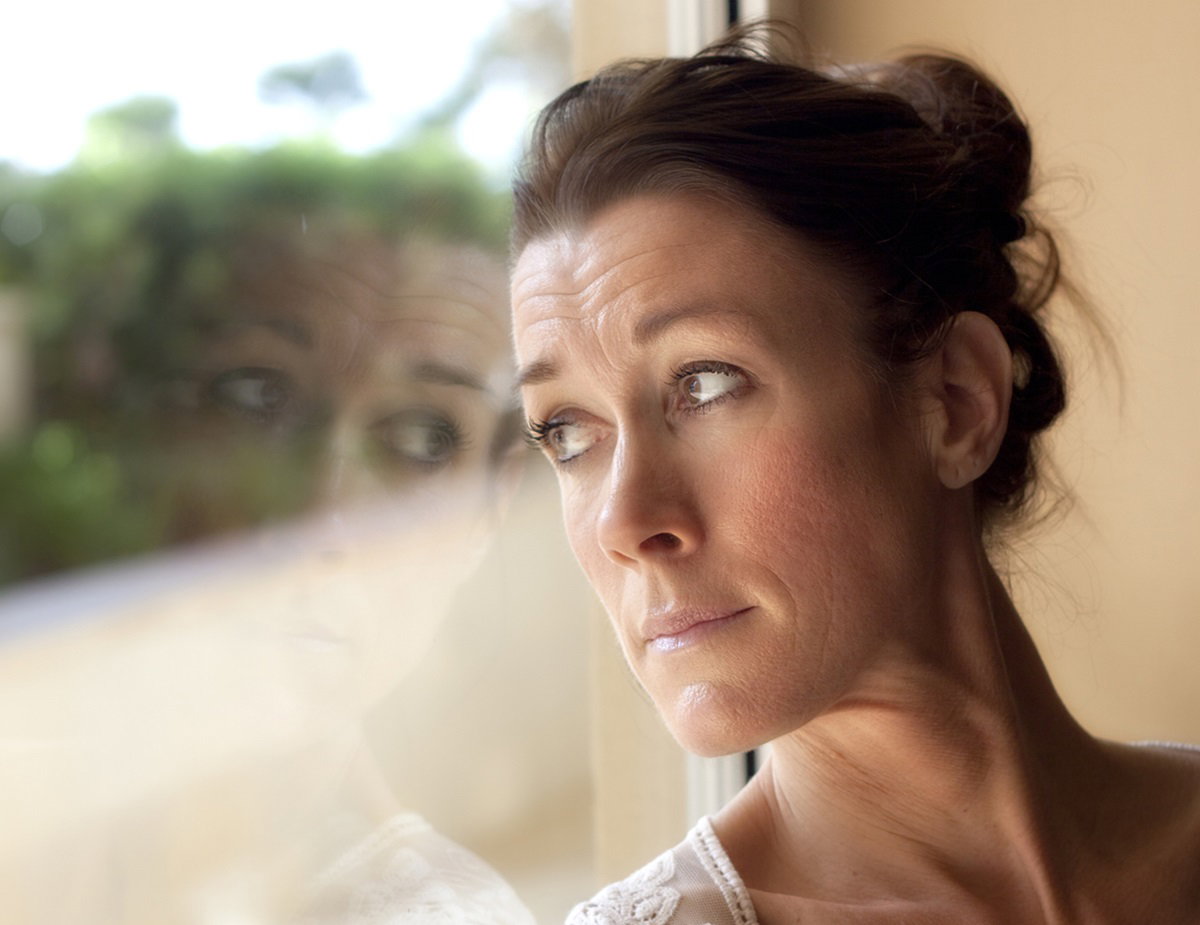 kobieta w średnim wieku z włosami spiętymi do góry patrzy zamyślona w okno