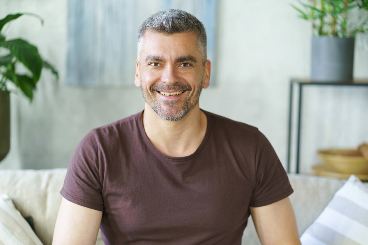 mężczyzna w średnim wieku z siwiejącymi włosami i zarostem siedzi na kanapie w salonie i szeroko się uśmiecha