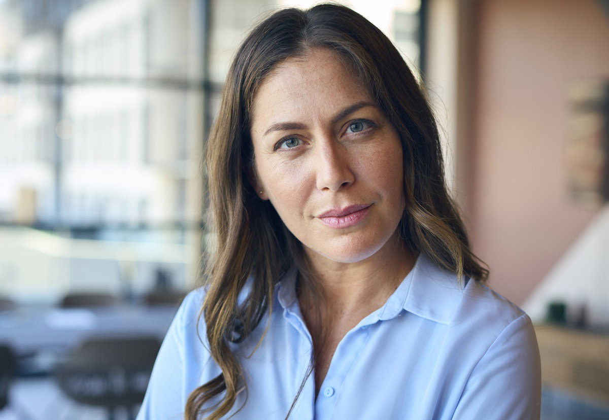 kobieta w średnim wieku z długimi włosami i jasnoniebieskiej koszuli stoi w pracy i patrzy poważnym wzrokiem