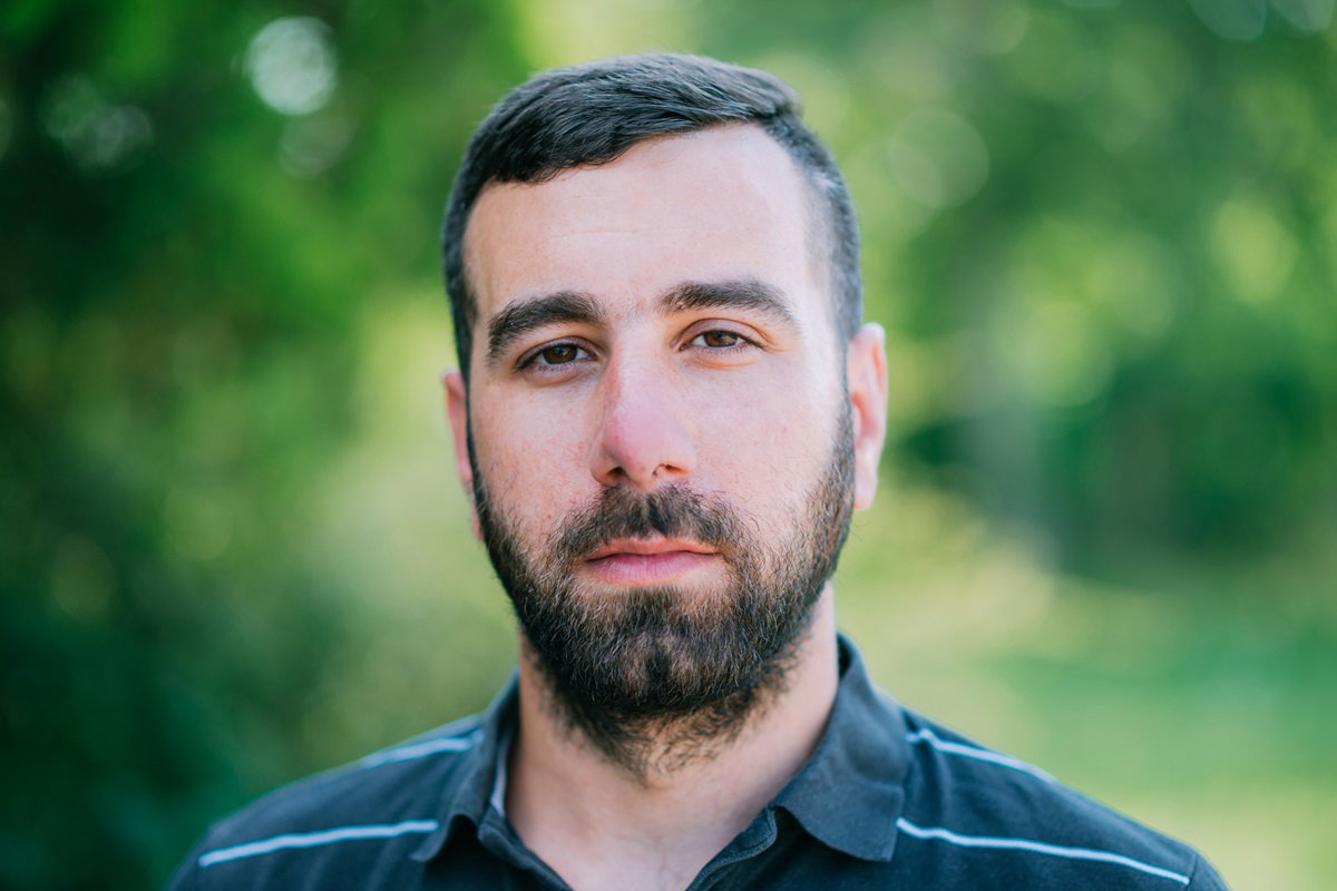 młody mężczyzna z wąsami i brodą stoi na łonie natury i patrzy poważnym wzrokiem