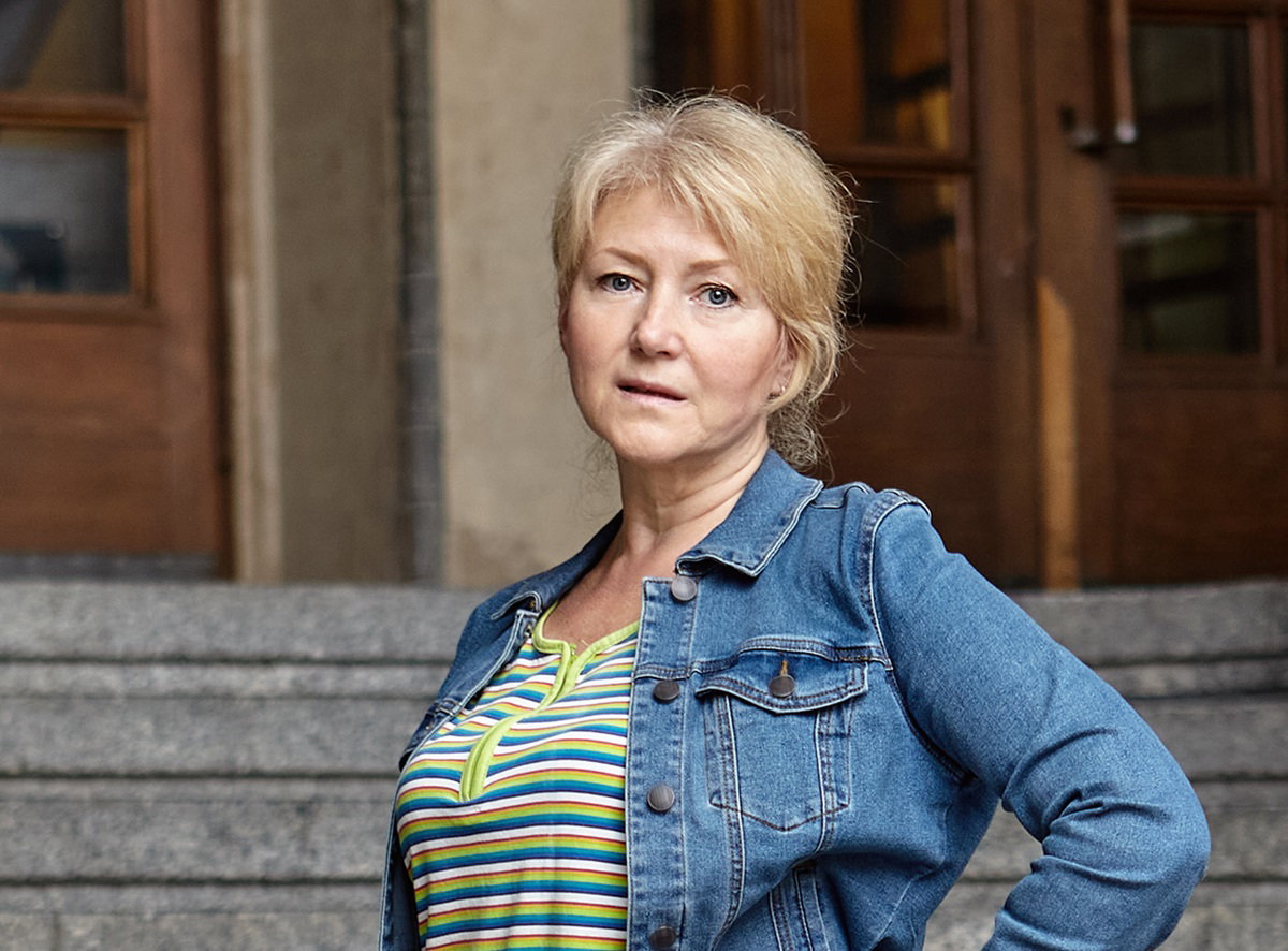 kobieta w dojrzałym wieku z blond włosami stoi na schodach na tle drewnianych drzwi i patrzy zaskoczonym wzrokiem