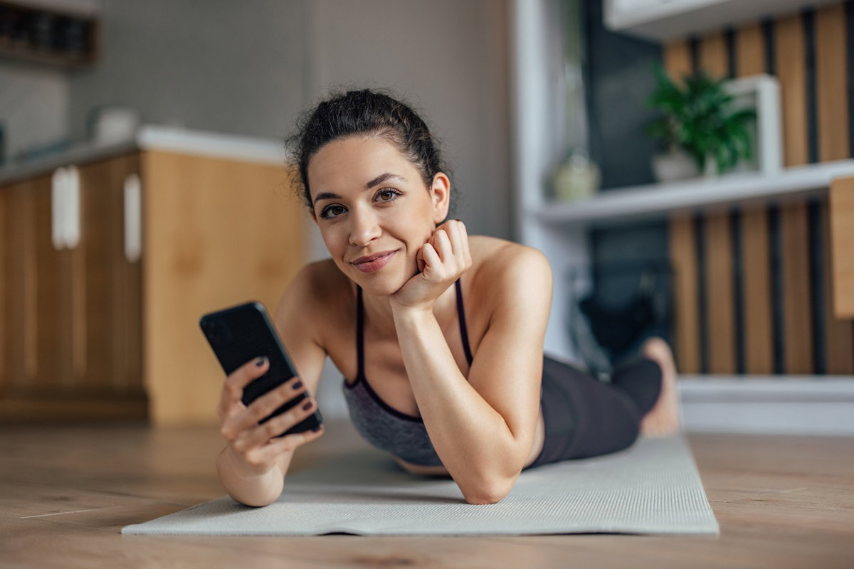 młoda dziewczyna leży na macie co ćwiczeń i trzyma telefon w ręce patrząc przed siebie z lekkim uśmiechem