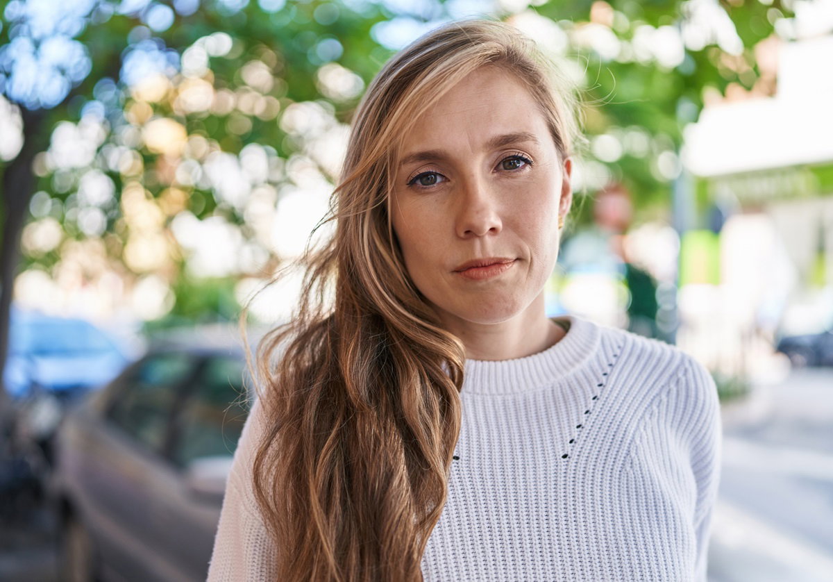 młoda kobieta z blond długimi włosami w białym swetrze stoi na tle zaparkowanych samochodów ze smutną miną