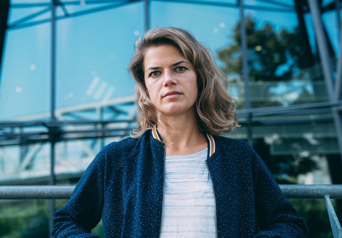 młoda kobieta z lekko falowanymi włosami stoi na tle szklanej konstrukcji i patrzy z politowaniem