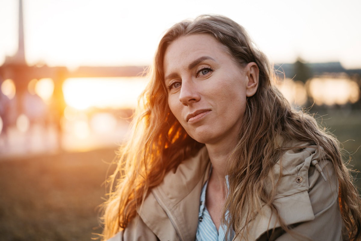 kobieta z długimi pofalowanymi blond włosami nad jeziorem patrzy sceptycznie przed siebie