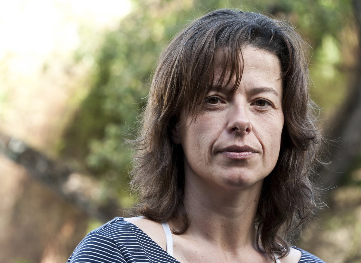 kobieta w średnim wieku z lekko kręconymi włosami stoi na dworze i patrzy poważnym wzrokiem