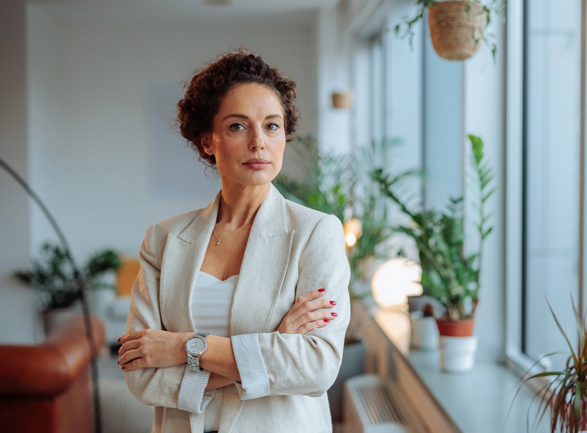 kobieta w średnim wieku z kręconymi upiętymi włosami  i w jasnej marynarce stoi ze skrzyżowanymi rękami i patrzy poważnym wzrokiem
